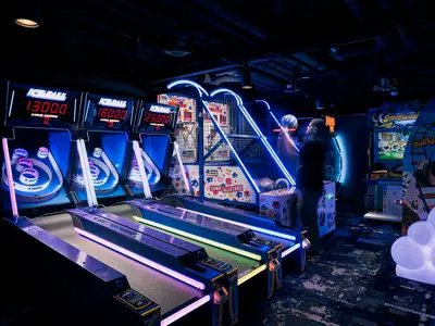 Ein lebhafter Arcade-Bereich an Bord der Norwegian Encore mit mehreren Skee-Ball-Automaten und einem Basketball-Spiel. Neonlichter tauchen den Raum in Blau- und Violetttöne, während eine Person am Basketballkorb steht.