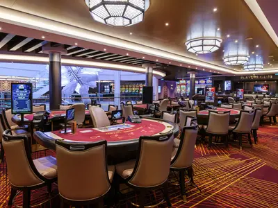 Ein weiter Blick in das stilvolle Casino an Bord der Norwegian Encore. Rote Spieltische und moderne Slotmaschinen füllen den elegant beleuchteten Raum, der menschenleer ist. Im Hintergrund ein Atrium.