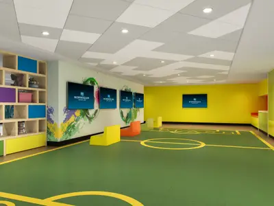 Der lebhafte Kinderbereich Splash Academy an Bord der Norwegian Encore: grüner Boden mit Spielfeldmarkierungen, bunte Wände, Regale mit Spielzeug und Bildschirme mit NCL-Logo.