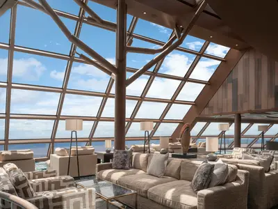 Die helle Haven Observatory Lounge an Bord der Norwegian Encore. Ein grosser Raum mit bequemen Sofas und Stühlen, hohen Fenstern mit Meerblick und einem Baum-ähnlichen Pfeiler unter einem Glasdach.