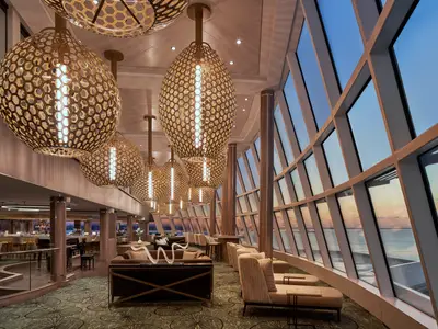 Die elegante Observation Lounge an Bord der Norwegian Encore zeigt stilvolle Sitzbereiche und dekorative Hängelampen. Grosse Fenster bieten Panoramablick auf das Meer bei Sonnenuntergang.