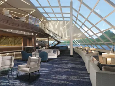 Blick in die zweistoeckige Haven Observation Lounge der Norwegian Encore mit riesigen Fensterfronten, die das Meer und gruene Berge zeigen. Helle, moderne Moebel und eine elegante Wendeltreppe praegen den Raum.