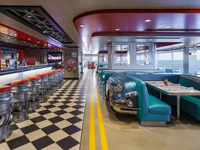 Innenansicht des American Diners an Bord der Norwegian Encore, gestaltet im Stil der 50er Jahre. Man sieht eine Bar mit Hockern aus Reifenfelgen, eine schwarz-weiss karierte Fliese und Sitzbänke in Türkisblau, die Autoteilen nachempfunden sind. Große Fenster bieten einen weiten Blick auf das Meer.