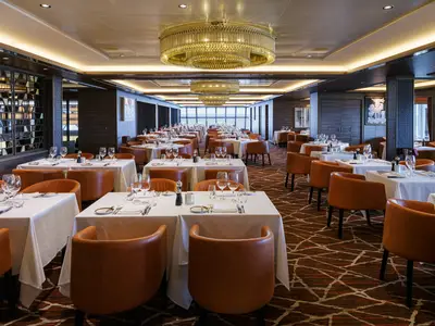 Blick in das stilvolle Steakhouse Cagney's an Bord der Norwegian Encore, das mit zahlreichen Tischen, braunen Ledersesseln und opulenten Kronleuchtern ausgestattet ist.