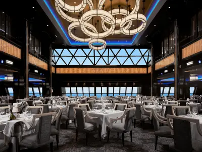 Eleganter Speisesaal "Manhattan Room" an Bord der Norwegian Encore. Zahlreiche gedeckte Tische und moderne Stühle mit Panoramablick auf das Meer durch große Fenster.