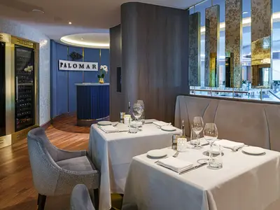 Elegantes Palomar Restaurant an Bord der Norwegian Encore. Gedeckte Tische, ein Empfangsbereich in Blau und ein grosser Weinkühlschrank. Moderne Einrichtung mit Holz- und Spiegelelementen.