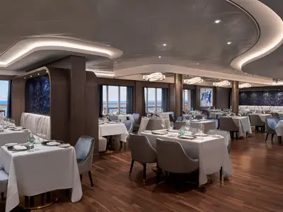 Innenansicht des modernen Palomar Restaurants an Bord der Norwegian Encore mit weiß gedeckten Tischen, eleganten Stühlen, Holzwänden und einem weiten Meerblick aus den Fenstern.
