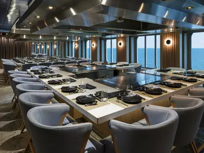 Das elegante Teppanyaki Restaurant an Bord der Norwegian Encore. Lange Tische mit Kochplatten und grauen Stühlen. Meerblick durch große Fenster, schwarzes Geschirr.