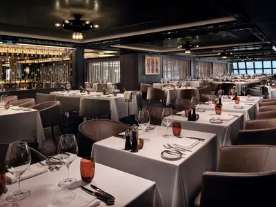 Elegantes Cagney's Steakhouse an Bord der Norwegian Encore. Zahlreiche Tische mit weissen Tischdecken, Gläsern und braunen Lederstühlen. Grosse Fenster im Hintergrund zeigen den Meerblick.