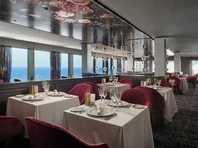 Ein elegantes Restaurant an Bord der Norwegian Encore mit dunkelroten Samtstühlen und weiss gedeckten Tischen. Grosse Fenster bieten einen weiten Blick auf das offene Meer.