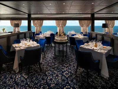 Blick in ein stilvolles Spezialitätenrestaurant an Bord der Norwegian Encore mit Tischen, die mit weissen Decken und Geschirr gedeckt sind. Blaue Samtsessel stehen an den Tischen. Grosse Fenster bieten Panoramablick auf das offene Meer.