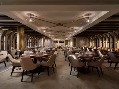 Innenansicht des eleganten Spezialitätenrestaurants Onda by Scarpetta an Bord der Norwegian Encore. Viele Tische mit Gedeck, moderne Einrichtung mit Holzvertäfelung und Goldakzenten, ohne Personen.