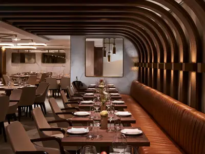 Das elegant eingerichtete Spezialitätenrestaurant Onda by Scarpetta an Bord der Norwegian Encore. Ein langer Tisch mit gepolsterter Bank im Vordergrund und weitere Tische mit bequemen Stühlen im Hintergrund. Die Wand ist mit Holzleisten und indirekter Beleuchtung gestaltet.