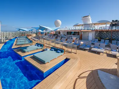 Weitwinkelaufnahme des sonnigen Oberdecks mit einem geschwungenen Pool, modernen Daybeds, Liegestühlen und einer Bar an Bord der Norwegian Encore. Im Hintergrund ist die Skyline zu sehen.