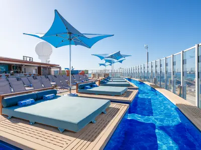 Das sonnige Sonnendeck Spice H2O der Norwegian Encore mit einem langen, flachen Pool, blauen Loungebetten, Sonnenschirmen und einer Bar. Im Hintergrund die Hafen-Skyline.