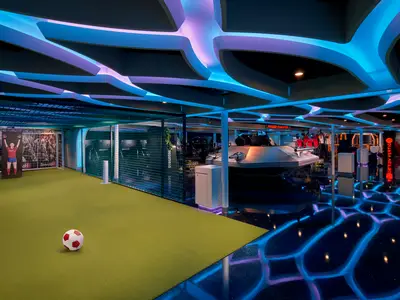 Sport- und Unterhaltungsbereich der Norwegian Encore. Links Fußballfeld mit Ball, rechts VR-Gaming-Stationen. Futuristische, blau-lila Neonbeleuchtung. Keine Personen.