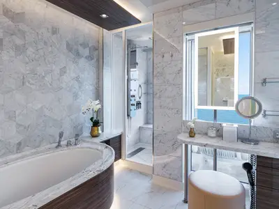 Helles, luxuriöses Marmor-Badezimmer einer Suite an Bord der Norwegian Encore. Mit ovaler Badewanne, begehbarer Dusche und Schminktisch mit Meerblick.