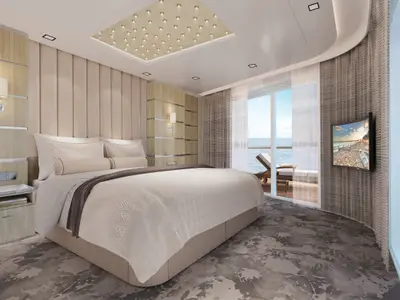 Blick in eine Balkonsuite der Norwegian Encore. Ein Doppelbett mit heller Bettwäsche, Nachttisch und Lampe. Ein Fernseher hängt an der Wand. Balkon mit Liegestuhl und Meerblick durch Glastür.