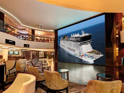 Ein weitwinkeliges Foto des eleganten, mehrstöckigen Atriums an Bord der Norwegian Epic mit Reihen von Sesseln und einem riesigen Bildschirm, der das Schiff auf offener See zeigt.