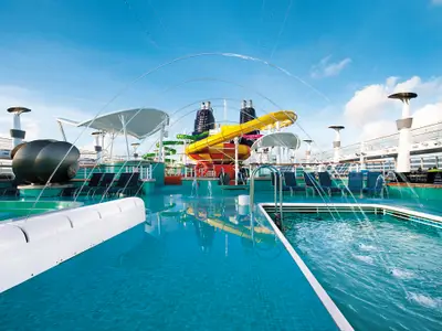 Wasserpark der Norwegian Epic: Grosser Pool mit Fontänen und Liegestühlen. Im Hintergrund bunte Wasserrutschen und Schornsteine des Schiffs unter blauem Himmel.