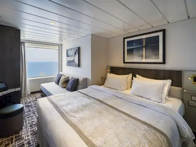 Eine geräumige Aussenkabine an Bord der Norwegian Epic mit einem komfortablen Doppelbett, einem angrenzenden Sofa und einem grossen Fenster, das einen weiten Blick auf das offene Meer bietet. Zwei Sektgläser stehen bereit.