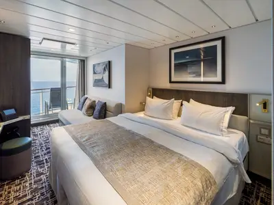 Eine helle und moderne Balkonkabine an Bord der Norwegian Epic. Ein großes Bett, ein Sofa und ein Balkon mit Stühlen bieten Meerblick über das offene Meer.