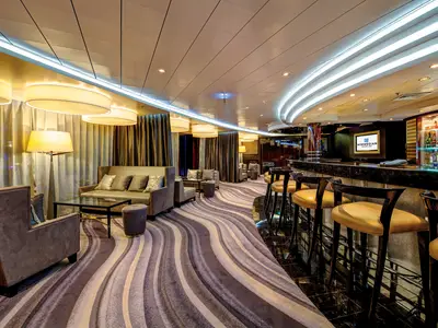 Die Bar-Lounge an Bord der Norwegian Epic. Sie zeigt eine geschwungene Bar mit Hockern und Sitzgruppen mit Sofas und Sesseln. Ein wellenförmiger Teppich und moderne Lichtinstallationen prägen das Ambiente.