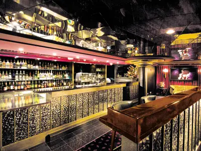 Elegante Bar-Lounge an Bord der Norwegian Epic mit einer gut gefüllten Theke, Spiegeldecke und gemütlichen Sitzbereichen. Ein großer Bildschirm zeigt ein Bild.