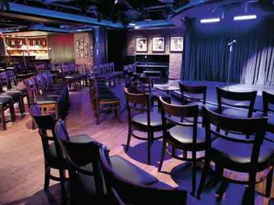 Ein leerer Comedy-Club an Bord der Norwegian Epic. Stuhlreihen sind auf eine Bühne mit Mikrofon ausgerichtet. Eine Bar ist sichtbar. Blaue und violette Beleuchtung schafft Abendstimmung.