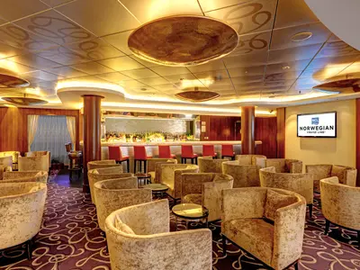 Shakers Bar auf der Norwegian Epic. Elegante goldene Sessel und Tische. Eine lange, beleuchtete Bar mit roten Hockern und vielen Flaschen. Goldene Decke mit Mustern und großen Leuchten.