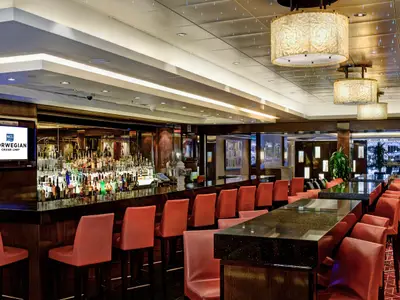 Elegante Maltings Bar an Bord der Norwegian Epic. Dunkle Theke, rote Barhocker, grosse Spirituosenauswahl. Stilvolle Hängelampen und Deckenbeleuchtung prägen den Raum.