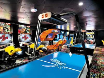 Die Spielhalle an Bord der Norwegian Epic mit Motorrad-Rennsimulatoren, Airhockey-Tisch und weiteren Arcade-Spielen. Der Raum ist dunkel, hat bunte Bildschirme und einen lebhaften Teppich. Ohne Personen.