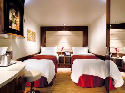Eine helle Innenkabine an Bord der Norwegian Epic mit zwei Einzelbetten, weisser Bettwäsche und roten Überwürfen. Ein Holzkopfteil, Nachttisch und eine Minibar sind sichtbar. Der Raum wirkt gemütlich und modern.
