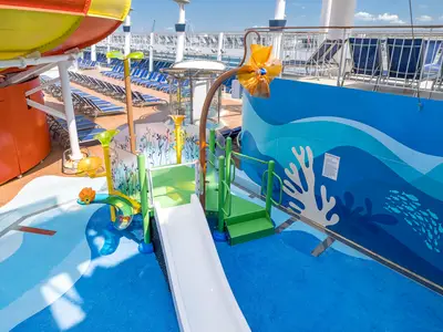 Blick von oben auf den bunten Kinder-Aqua-Park an Bord der Norwegian Epic. Eine weiße Rutsche führt ins flache Wasser, umgeben von Wasserspielen und einem farbenfrohen Wandbild.