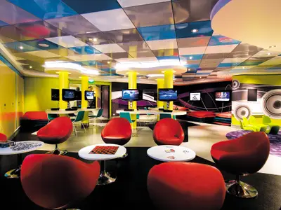 Ein farbenfroher, modern gestalteter Jugend- und Gaming-Bereich an Bord der Norwegian Epic. Rote Sessel um kleine Tische mit Spielen, gelbe Saeulen und zahlreiche Bildschirme mit Konsolen praegen den Raum.