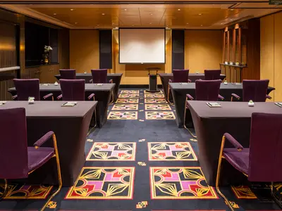 Eleganter Konferenzraum an Bord der Norwegian Epic. Reihen von Tischen und violetten Stühlen. Ein Rednerpult und eine Projektionsfläche im Hintergrund. Warme Holzvertäfelung.