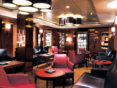 Die Humidor Cigar Lounge an Bord der Norwegian Epic. Ledersofas und Sessel in Schwarz und Rot stehen um Holztische. Holzvertäfelung und warme Beleuchtung schaffen eine elegante, gemütliche Atmosphäre.