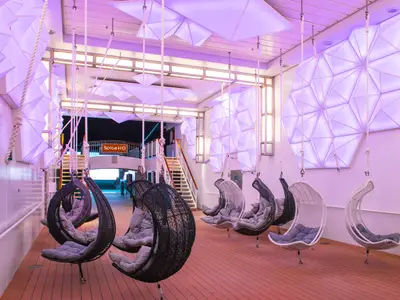 Lounge-Bereich 'Spice H2O' an Bord der Norwegian Epic. Moderne Hängesessel stehen auf einem Holzdeck. Wände und Decke sind mit violetten, geometrischen Lichtinstallationen gestaltet.