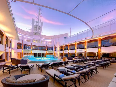 Abendliche Ansicht des The Haven Courtyard Poolbereichs an Bord der Norwegian Epic. Blaue Pools und Jacuzzi umgeben von Liegestühlen und Cabanas, beleuchtet von warmem Licht unter einem rosafarbenen Himmel.