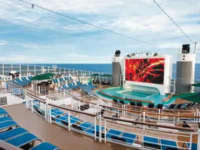 Weitwinkelaufnahme des lebhaften Pool- und Sonnendecks an Bord der Norwegian Epic mit vielen Liegestühlen und einem grossen LED-Bildschirm vor dem weiten blauen Meer und Himmel. Grüne Sonnenschirme spenden Schatten.