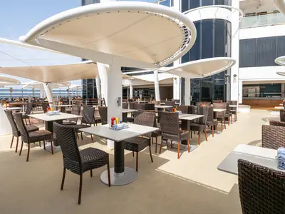 Blick auf das Aussenrestaurant Great Outdoors an Bord der Norwegian Epic. Leere Tische und Korbstühle unter hellen Sonnensegeln. Hinten das Meer und das Schiffsgebäude mit Glasfronten zu sehen.