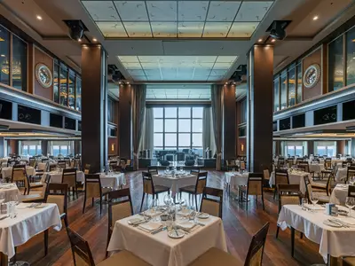 Der weitläufige Manhattan Room, ein elegantes Hauptrestaurant an Bord der Norwegian Epic, ist mit vielen gedeckten Tischen ausgestattet. Im Hintergrund befindet sich eine Bühne mit Instrumenten vor großen Fenstern, die Meerblick bieten. Braune Säulen und Holzböden prägen den Raum.