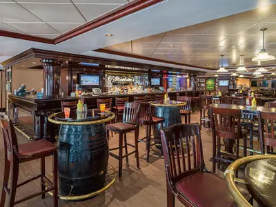 Einblick in O'Sheehan's Bar & Grill an Bord der Norwegian Epic. Lange dunkle Holzbar mit Flaschen, Bildschirmen und Hockern. Viele Holztische, einige auf Fässern, mit Stühlen und Getränken. Eine gemütliche Pub-Atmosphäre ohne Gäste.