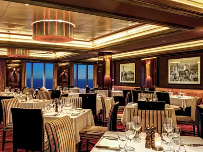 Elegantes Spezialitätenrestaurant Cagney's Steakhouse auf der Norwegian Epic. Gedeckte Tische, gestreifte Stühle, Abendmeerblick. Luxuriöses Holzinterieur mit Kunstwerken.