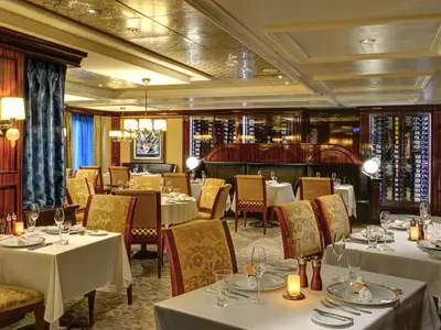 An elegant dining room, Le Bistro, on the Norwegian Epic. Tables are set with white cloths, surrounded by patterned chairs. Dark wood walls, chandeliers, and illuminated wine racks are visible.