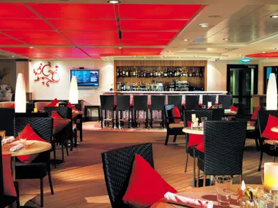 Moderne Bar-Lounge an Bord der Norwegian Epic. Rote Deckenplatten und Sitzkissen prägen den Raum. Man sieht Tische, Stühle, Barhocker und eine voll ausgestattete Bar mit TV-Bildschirm.