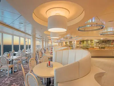Modernes Garden Café Buffet-Restaurant an Bord der Norwegian Epic. Helle Sitzbereiche, grosse Fenster mit Meerblick bei Sonnenuntergang und Buffetauslagen sind sichtbar.