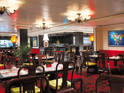 Restaurant Shanghai's auf der Norwegian Epic. Dunkle Holzmöbel mit roten Sitzpolstern und Teppich. Beleuchtet von Kronleuchtern und Laternen. Im Hintergrund eine offene Küche.