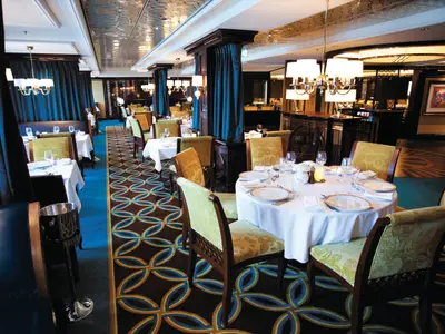 Blick in ein stilvolles Restaurant an Bord der Norwegian Epic. Gedeckte Tische, goldene Sessel und ein Teppich mit blau-goldenem Muster sind zu sehen.