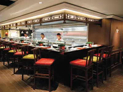 Gastronomisches Erlebnis in der Noodle Bar an Bord der Norwegian Epic. Zwei Crewmitglieder bereiten Gerichte hinter einer Bar mit Sitzplätzen zu. Moderne Einrichtung mit Holz und roten sowie grünen Polstern.